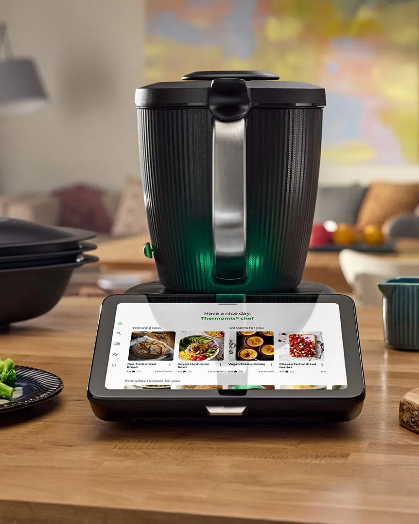 Thermomix® TM7 auf einer Holztheke in einer hellen Wohnküche, daneben ein Teller mit Lachs und Gemüse